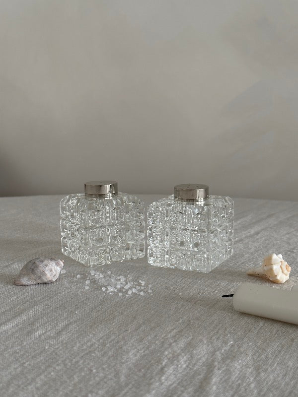 Two clear glass containers with silver lids on a textured surface with shells and a candle.