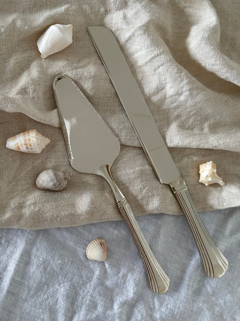 Silver cake server and knife set on a textured fabric surface with seashells.